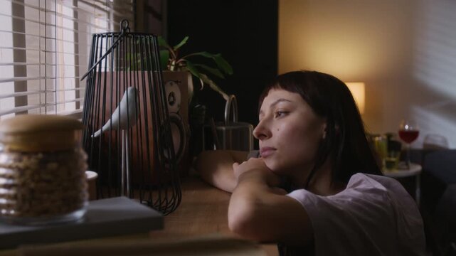 Calm young woman resting her head on her hands, leaning on windowstill and looking out while being under house arrest