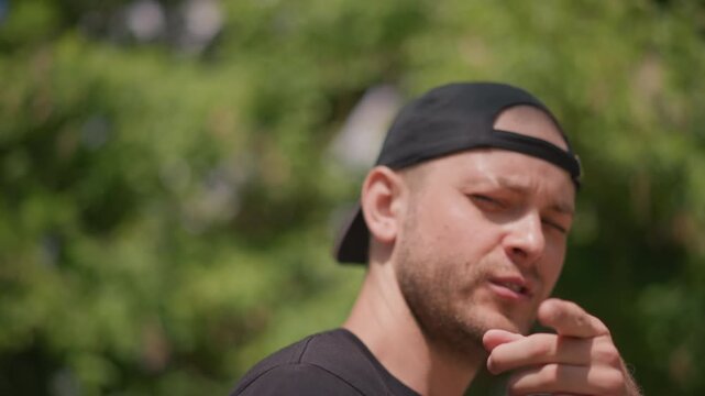 White Man Outdoors Looking Upward With Attitude, Street Performer And Coach Gestures, Confident Stare, Cap Backwards, Summer Trees Bokeh, Expressive Jaw Movements, Candid Portrait, Swaggering Urban