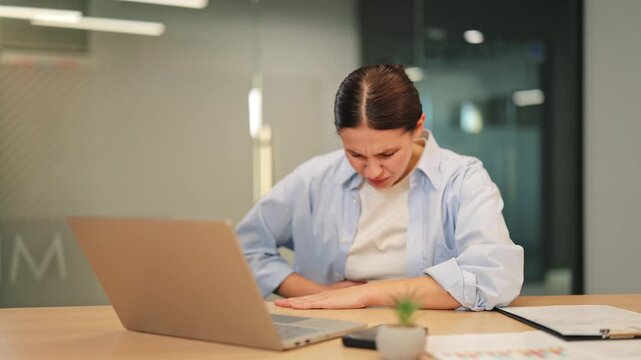 Woman experiences severe stomach pain while working at office desk. Discomfort causes distress, impacting productivity. Feeling ill and suffering, woman hopes relief comes soon from abdominal ache.