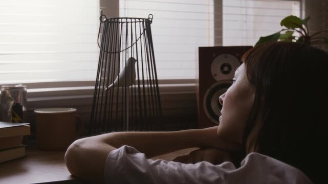 Over the shoulder shot of bored young woman looking at toy bird in cage while staying at home during house arrest