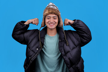 Confident young Asian man flexing muscles in winter clothing showing he is strong on blue backdrop