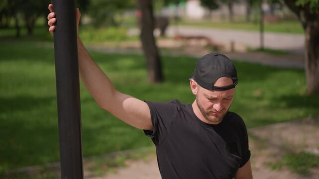 man stretching arm on pole, outdoor warmup and recovery scene showing man leaning against street pole, extending arm across body with slight squint from sun, casual cap and tshirt, leafy path