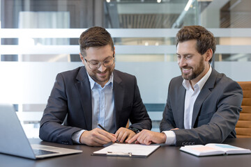 Two businessmen smiling and signing a contract at a modern office meeting table, collaborating on a formal agreement and partnership with paperwork, pens and laptops nearby