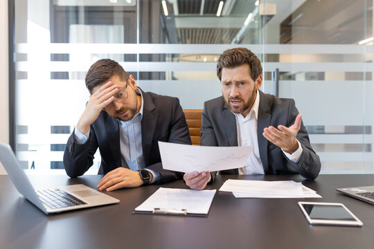 Two stressed businessmen sitting at an office table, discussing a financial report showing negative results, one holding his head in despair while the other explains the problem with concern