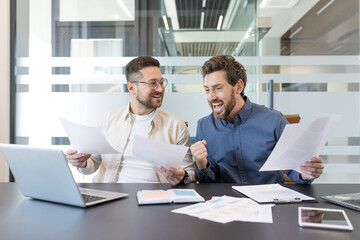 Two overjoyed businessmen celebrating an achievement, fist pumping and smiling while holding...