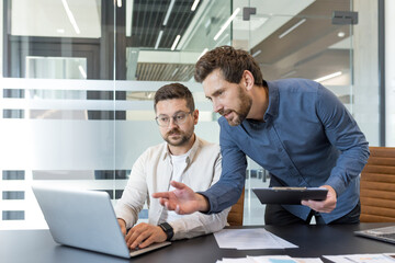 Two businessmen work together analyzing data on a laptop in a bright modern office, demonstrating teamwork, collaboration, and professional focus during a business meeting