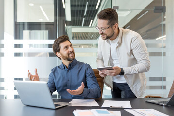 Two business partners are discussing ideas and strategy, smiling and collaborating on a project with a laptop and digital tablet in a modern office environment