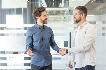 Two smiling adult businessmen standing in a modern office, shaking hands and discussing a...
