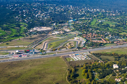 Aerial view of a racetrack snaking through the sunlit landscape, bordered by lush greenery and the distant cityscape, a vibrant contrast of speed and serenity, Madrid, Community of Madrid, Spain.