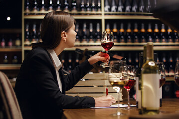 Experienced female sommelier tasting wine into glass sitting in cellar over plenty bottle shelf background.