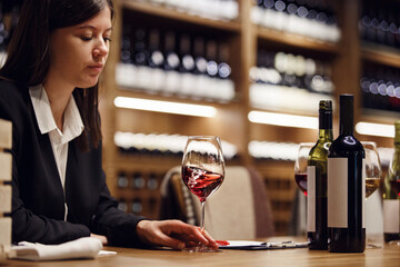 Female sommelier swirling red wine in glass on cellar background being in cellar. Wine expert exam to study different wine. 