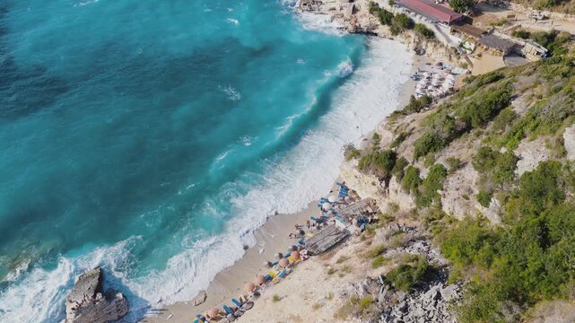 Aerial view of turquoise waters meet sandy shores, dotted with colorful umbrellas, creating a vibrant contrast against the lush greenery, Ksamil, Qarku i Vlores, Albania.