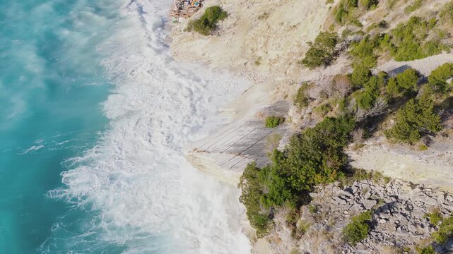 Aerial view of crashing waves meeting a rugged shoreline, the turquoise waters contrasting with the stark cliffs and verdant vegetation, Ksamil, Qarku i Vlores, Albania.