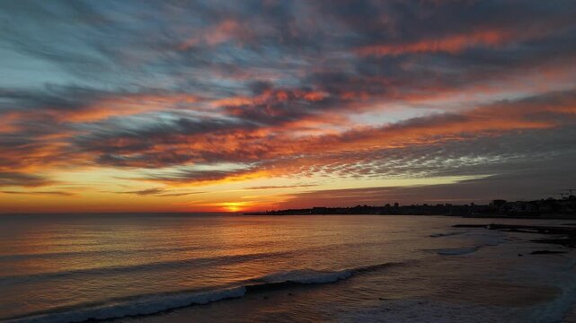 Aerial view of ocean waves gently lapping the shore under a vibrant sunset, painting the sky with hues of orange and blue, Estoril, Lisbon, Portugal.