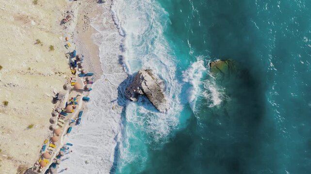 Aerial view of turquoise waves crashing against rocky outcrops and sandy beach dotted with umbrellas, creating a scene of coastal bliss, Ksamil, Qarku i Vlores, Albania.