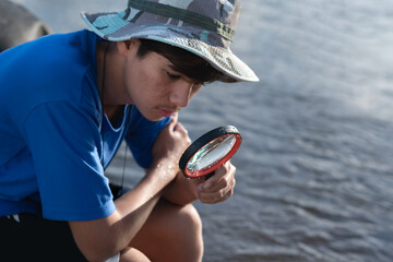 A boy sitting and using magnifying glasses at river, exploring tiny life forms contained within a clear plastic bottle. Igniting curiosity and the spirit of scientific discovery.