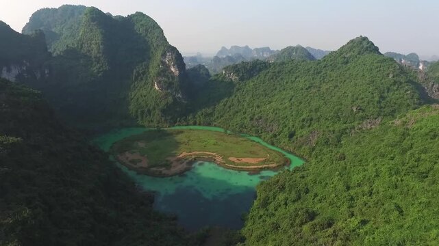 Aerial view of Trang An Valley in Vietnam: A vibrant turquoise lake is nestled between majestic, lushly forested limestone cliffs under a clear, sunny sky, showcasing iconic karst scenery.