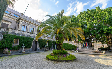 CATANIA, ITALY. The lush courtyard of Villa Cerami, a historic 18th-century palace currently housing the Faculty of Law, featuring a large palm tree, stone statues, and a grand stairc