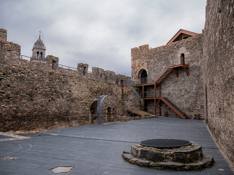 View of an interior courtyard within the walls of the Templar castle of Ponferrada with a well on one side and a wooden staircase leading to different rooms, and the tower of the Basilica of the Virgi