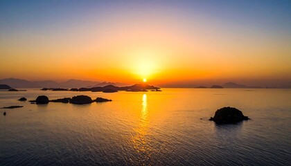 Fototapeta premium Aerial view captures a serene sunset over an archipelago. Vibrant orange hues paint the sky and reflect on the calm ocean. Several islands dot the horizon
