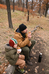 Fototapeta premium A father and son eat outdoors and drink tea from a thermos. An outdoor picnic in winter without snow or autumn