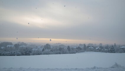 Fototapeta premium Showing large snowy field occupying center near distant houses and trees, with falling snowflakes
