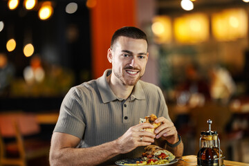 Young adult man smiling, eating a sandwich in a restaurant