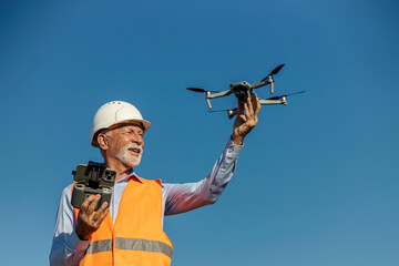 Senior male environmentalist wearing safety gear launching drone outdoors under clear blue sky