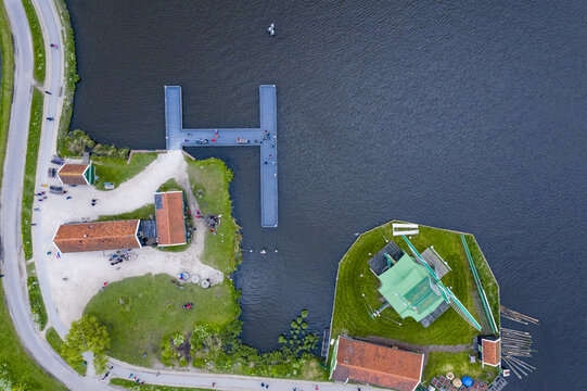 Aerial view of green-roofed windmills and red-tiled buildings meet the tranquil waters near a pier bustling with tiny figures, Zaandijk Zaanse Schans, Zaandam, Netherlands.