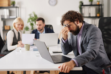 Fototapeta premium People are working in an office setting, with one person looking stressed and tired at a laptop while others are discussing papers and tasks at their desks.