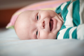 A baby wearing a striped green and white shirt is lying on their back on a light blue bedspread. The baby is smiling with their mouth open and looking at the camera.
