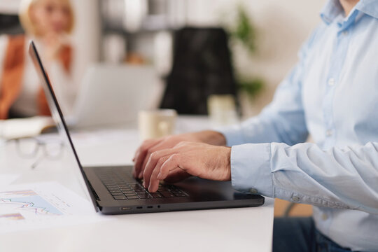 A person sits at a desk in a shared office space. They are focused on typing on a laptop. Other people can be seen in the background working quietly.