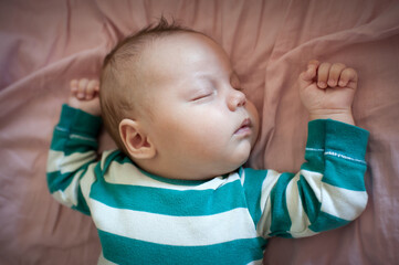Small child wearing a striped green and white shirt rests peacefully on a pink sheet. Baby has closed eyes and a relaxed face while sleeping.