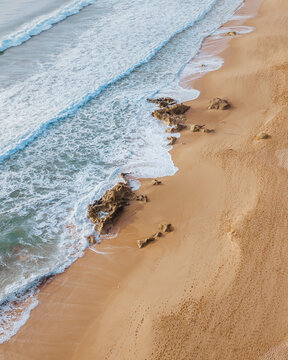 Aerial view of the shoreline where foamy waves crash against the sandy beach dotted with dark rocks, Algarve, Faro District, Portugal.
