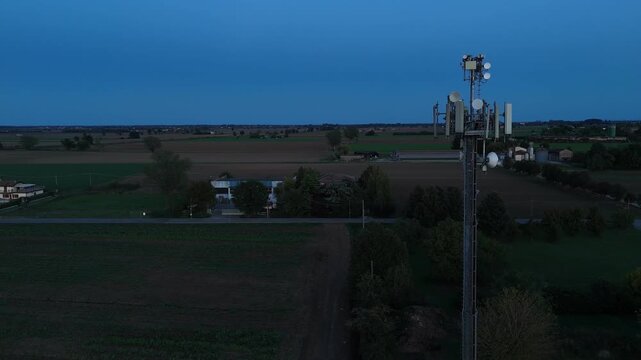Slow drone pan at blue hour shows a tall cell‑tower rising above the flat agricultural landscape of the Po Valley in northern Italy, multiple sector antennas and microwave dishes, digital connectivity