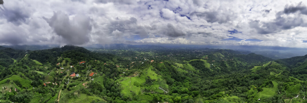 Aerial view of emerald hills blanketed in verdant foliage, punctuated by terracotta rooftops under a sky streaked with silver clouds, Atenas, Costa Rica.