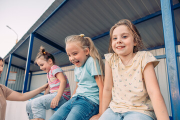 Three young girls sit together on a ledge, enjoying quality time. They have different hair colors and styles, wearing casual clothing. setting is bright and cheerful.
