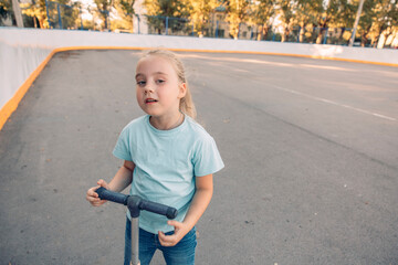 young girl rides scooter on a smooth surface. She has blond hair and wears a blue shirt. The scene captures outdoor activity.