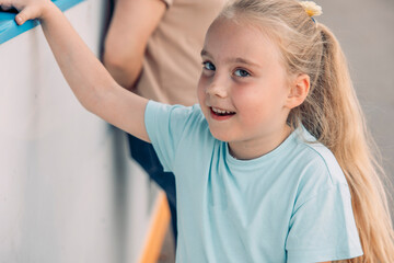Young Caucasian girl with long blonde hair smiles while leaning against a wall in an outdoor court setting. She enjoys quality time with friends.