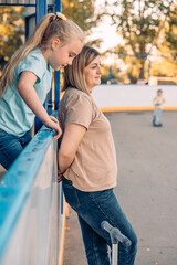 A young blond girl and a woman with brown hair enjoy quality time outdoors at a sports court. They lean over a railing, watching activities on the court.