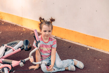 Young Caucasian girl with brown hair in pigtails sits on the ground next to a pink bicycle. She enjoys quality time outdoors on a court.