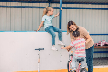 woman helps a young girl ride a pink bicycle. The girl has brown hair in a bun and wears a striped shirt. A boy climbs on a railing nearby. They enjoy quality family time.