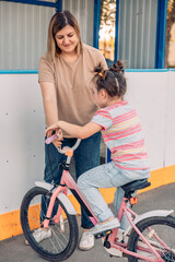 young girl rides a pink bicycle while her mother supports her. They are enjoying quality time together in a sunny outdoor basketball court, vertical