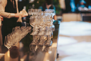 A bartender arranges empty glasses on a shiny bar counter at a lively event venue. Guests move...