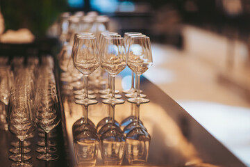 Empty wine glasses are arranged neatly on a shiny bar surface. The setting is inside a restaurant during the evening hours. Soft light reflects off the glasses and bar