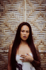 Confident woman in long printed dress standing in front of bamboo wall, intense expression and tropical editorial style