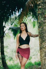 Barefoot woman standing in tropical jungle surrounded by palm trees, red wrap skirt and confident outdoor pose