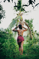 Back view of woman walking through tropical forest barefoot, red sarong and long hair in jungle path