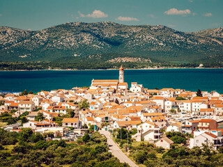 Aerial view of red-roofed buildings cluster around a towering church steeple by the tranquil sea, framed by distant mountains, Murter, Sibenik-Knin County, Croatia.