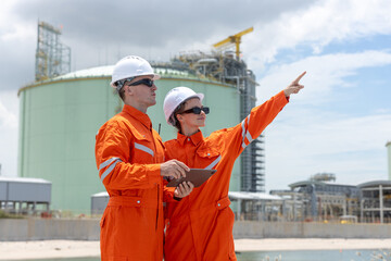 Engineers in orange safety uniforms and helmets reviewing project direction at an industrial site near water, representing teamwork, planning, infrastructure development in the energy sector.
